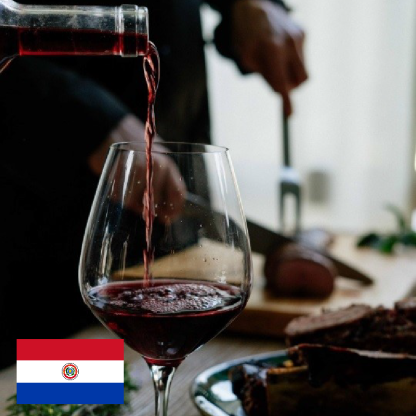 Fotografia. Taça de vinho meio cheia. Mais vinho está sendo servido na taça. Ao fundo, uma mesa com carne. No canto inferior esquerdo, a bandeira do Paraguai.